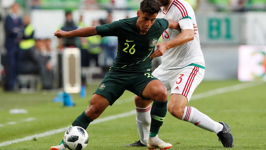 An athlete dressed in dark green maintains control of a soccer ball that an athlete in white wants