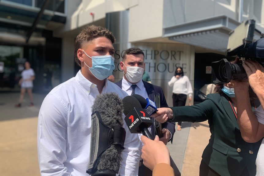 Man wearing mauve shirt and blue face mask fronts media microphones and cameras in front of court