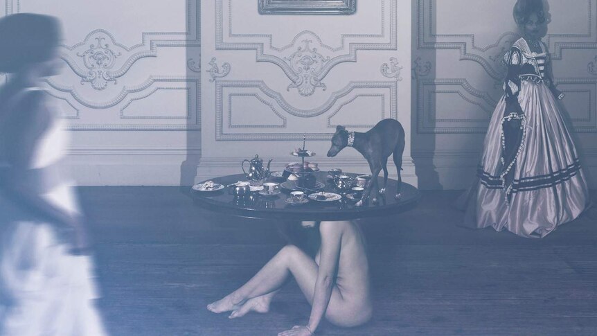 A naked women sits on the ground supporting a table, upon which rests a traditional afternoon tea and a dog