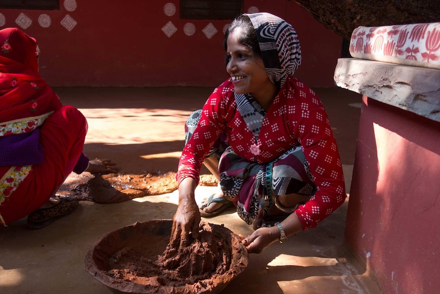 A woman in a red top crouches over a bowl and mixes natural colours.