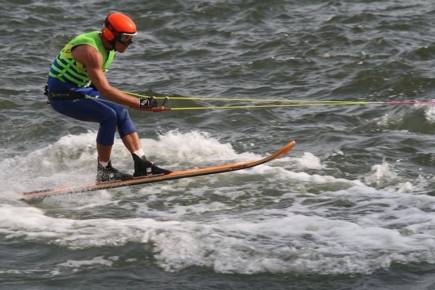 Water skier wears red helmet, yellow vest and blue leggings. Man on skis holding ropes.
