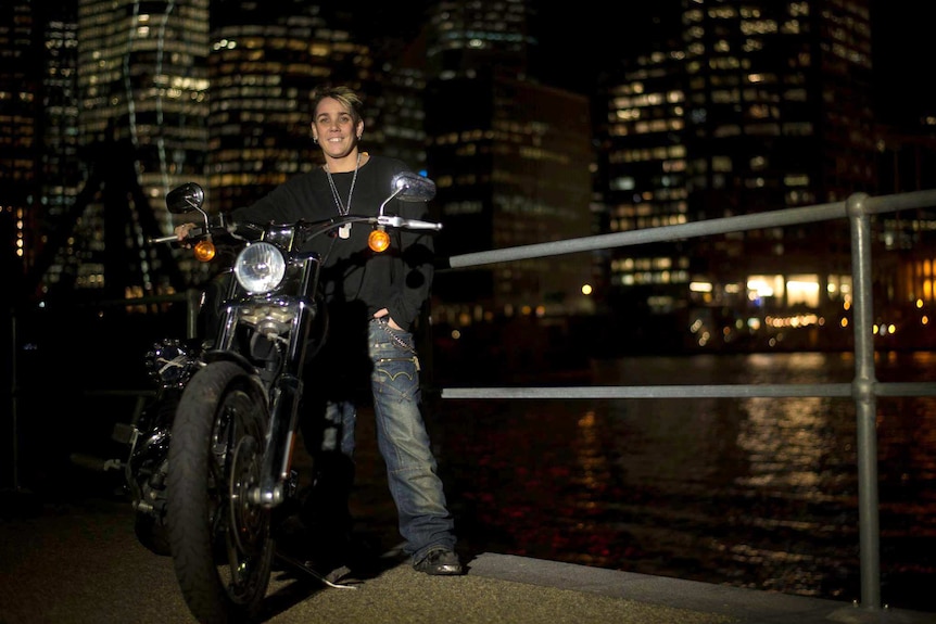 Julz Raven stands next to her bike with the city high rise lights in the background.