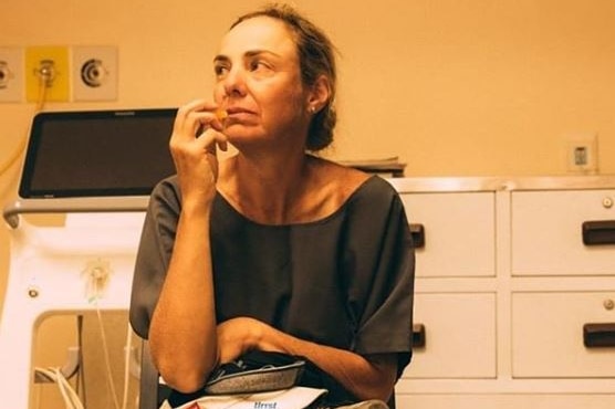 Mina Guli wears dark grey hospital gown as she sits on a chair wither a hat and shoes in lap with hospital equipment behind her.