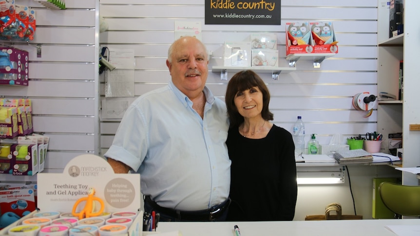 A man and a woman stand side by side smiling behind a shop counter with baby toys on shelves behind and in front of them.