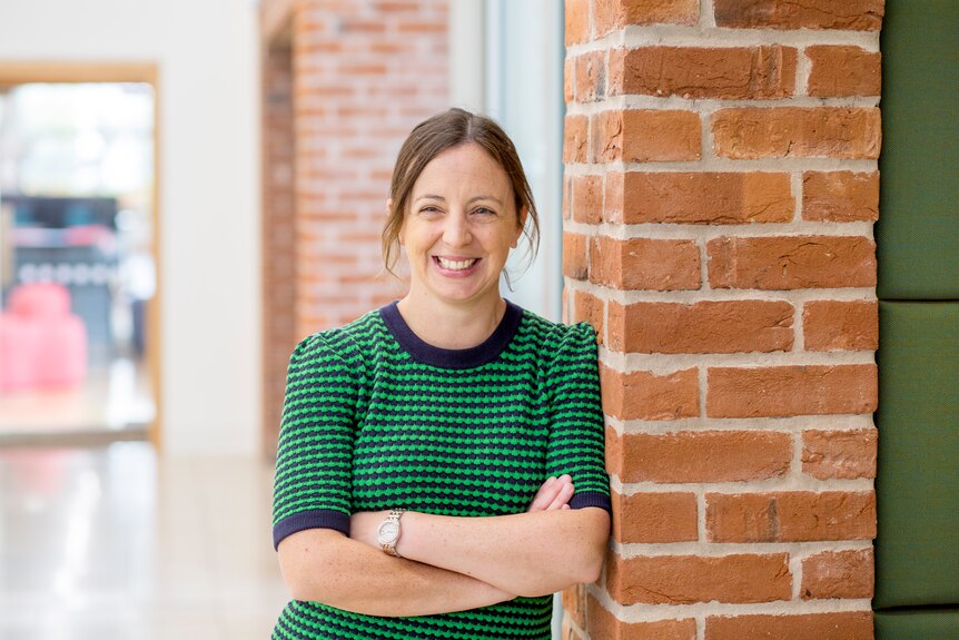 Profile picture of Professor Kirsty Elliott-Sale. She is smiling as she crosses her arms and leans on a brick wall.