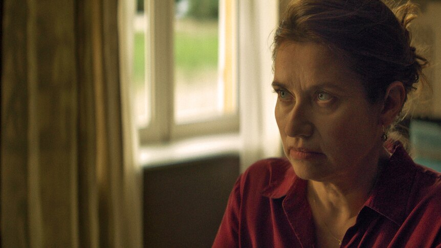 A woman in a red shirt sits in front of a window and stares into the distance 