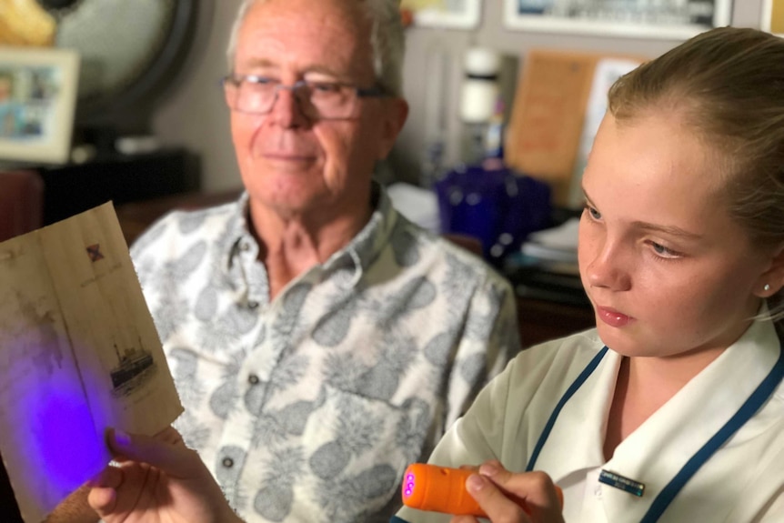 A girl in a school uniform holding a black light to an old piece of paper beside her grandfather.