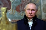 Vladimir Putin, wearing a white turtleneck jumper under a thick navy coat, looks sad