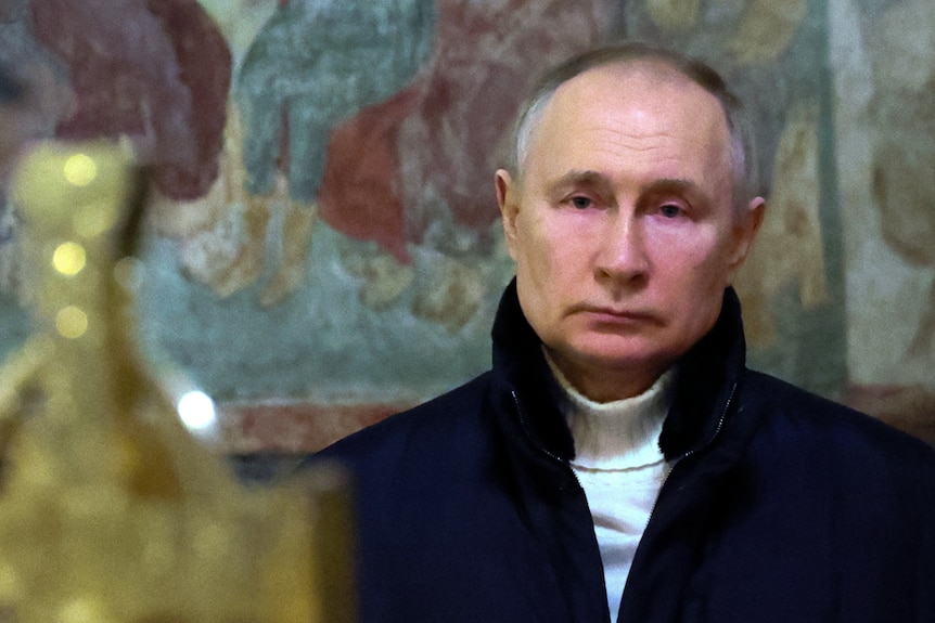 Vladimir Putin, wearing a white turtleneck jumper under a thick navy coat, looks sad