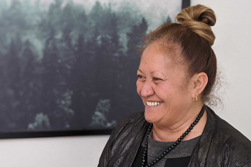 A woman wearing a black jacket and black bead necklace smiles at another woman. A large photo of a forest hangs behind her.