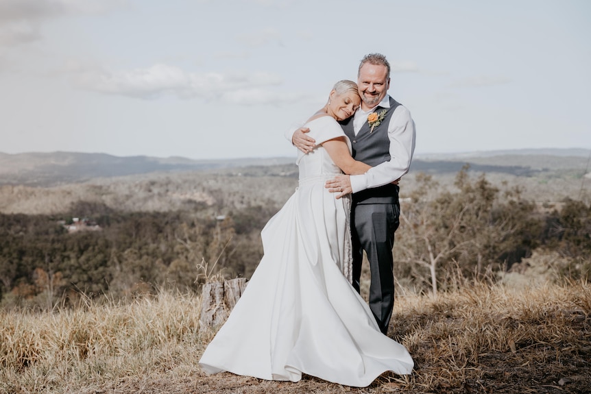 A wedding photo of the married couple holding each other on a hillside with a view over countryside.