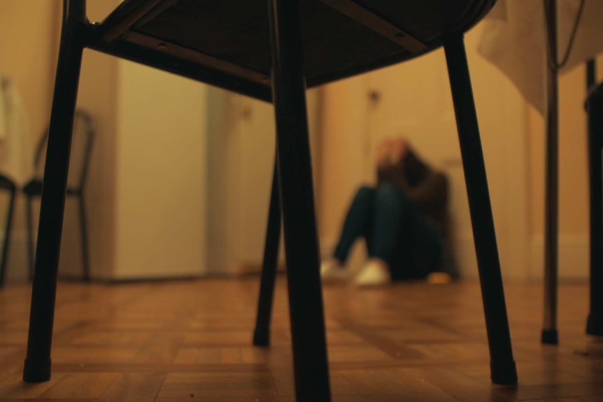 An unidentifiable but distraught child sits on the floor in a kitchen holding its head in its hands.