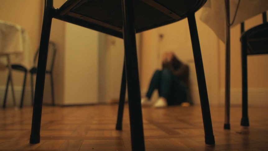 An unidentifiable but distraught child sits on the floor in a kitchen holding its head in its hands.