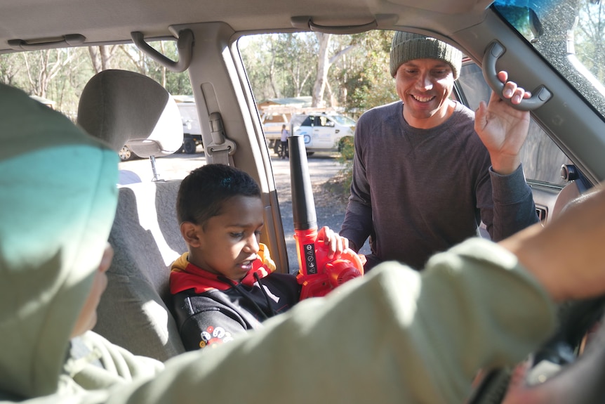 An Indigenous man in a grey sweater and a beanie leans into a car smiling at a child.