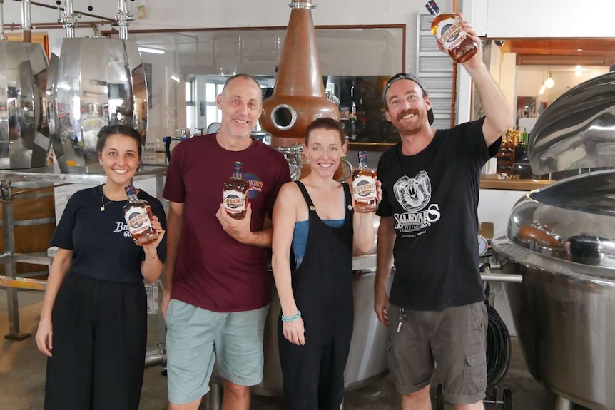 Two men and two women stand in a distillery holding bottles of rum, smiling and looking happy.