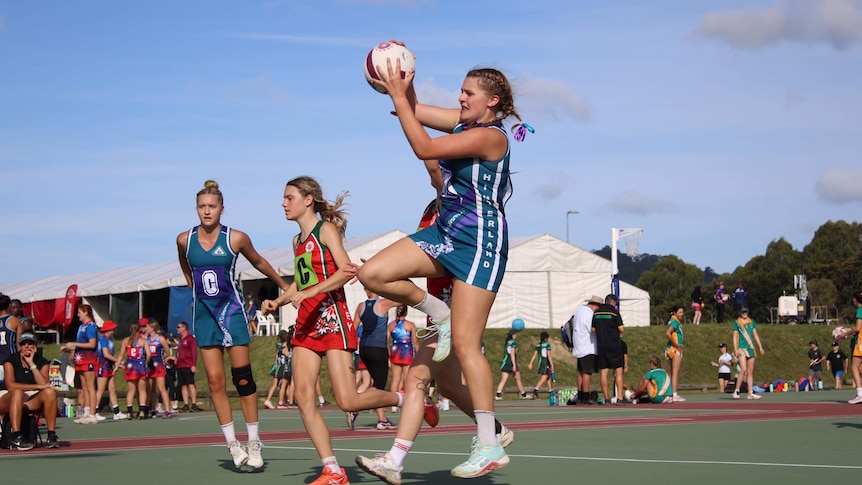 Young girls wearing red, blue netball uniforms playing netball as other chat on the sidelines or walk in front of a marquee. 