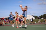 Young girls wearing red, blue netball uniforms playing netball as other chat on the sidelines or walk in front of a marquee. 