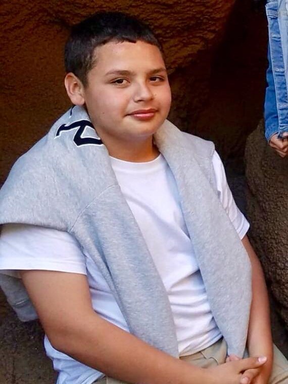 A young boy with Latino features dressed in a white t-shirt with a grey sweater pulled over his shoulders poses for a photo