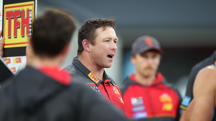 A man wearing a Gold Coast Suns jacket talks to a group of players.