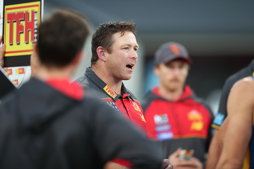 A man wearing a Gold Coast Suns jacket talks to a group of players.