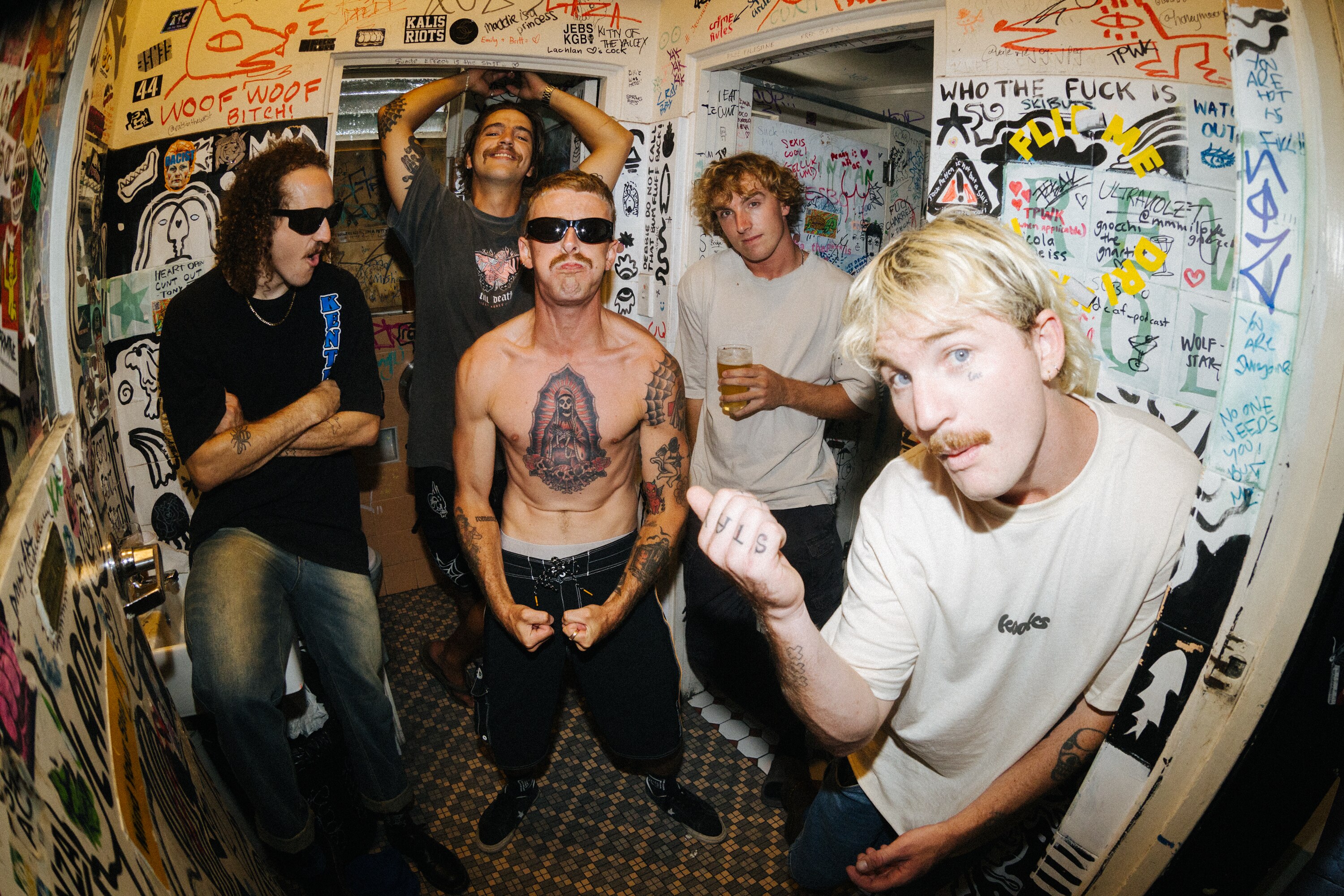 Five men stand together in a heavily-graffitied bathroom. Two are wearing sunglasses and one is shirtless.