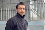 A young man stands in front of a grey building.