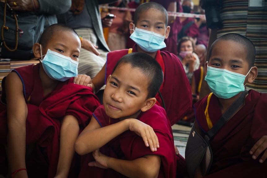 Four young boys in maroon robes, three wearing surgical masks, pose for the camera while sitting together outside