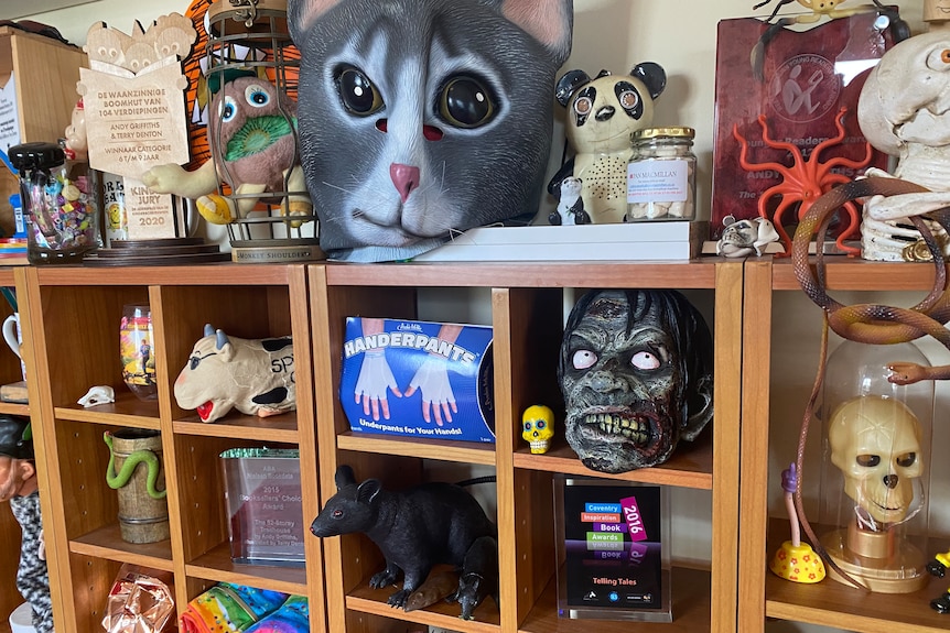 A bookcase filled with strange objects including a cat mask, panda toy, zombie head, large rat toy