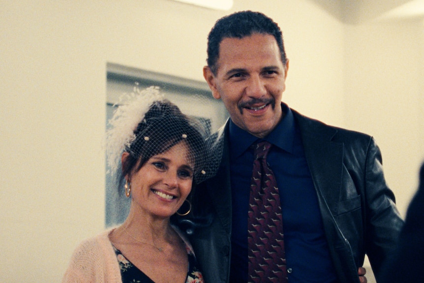 A middle-aged white woman wearing a bridal veil smiles while standing next to a middle-aged white man in a black leather jacket.