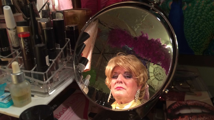 A man wearing a wig looks at himself in the mirror