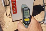 A person holds a handheld thermometer with a reading of 50.5, in front of a fuel pump.
