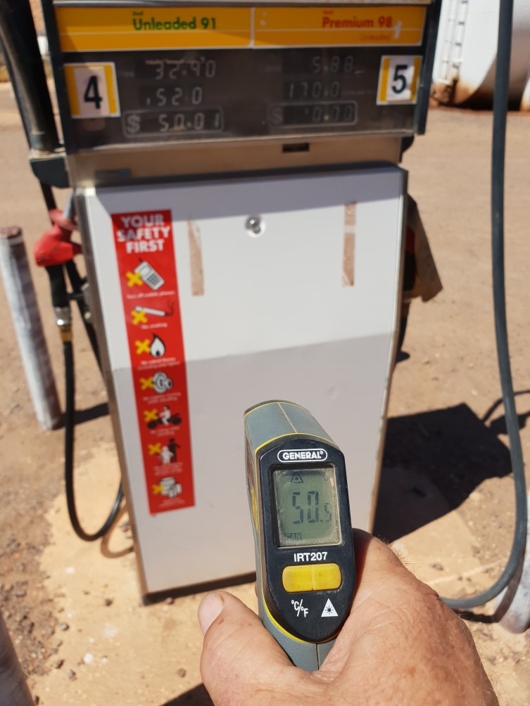 A person holds a handheld thermometer with a reading of 50.5, in front of a fuel pump.