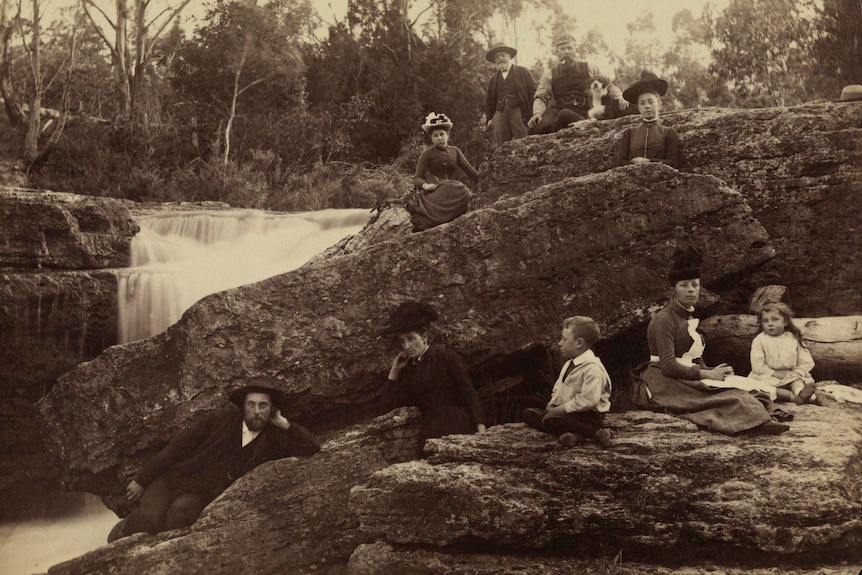 a black and white photo from the 1800s of people sitting around on rocks at a rivulet, wearing clothes from the olden days
