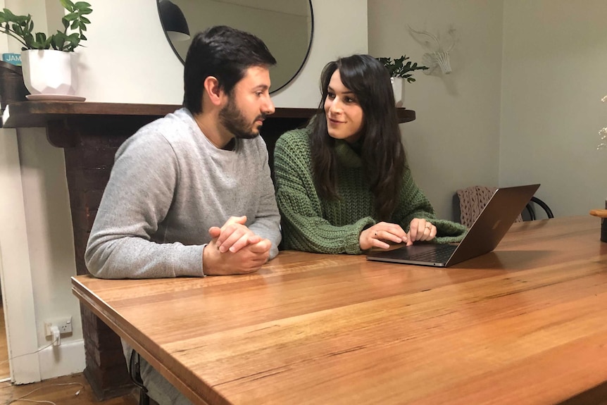 A dark-haired young couple sit at a table in front of a laptop computer