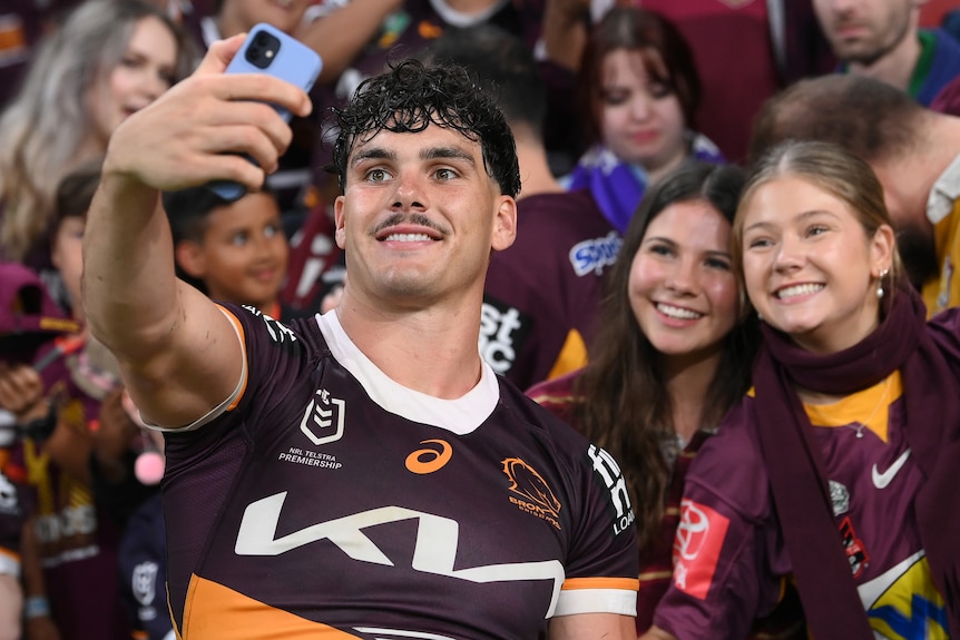 A Brisbane Broncos player smiles as he takes a picture with his mobile phone, while two young female fans smile behind him.