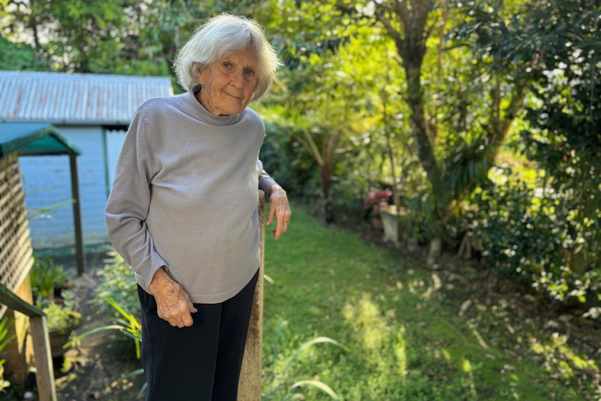 An older woman with white hair stands on a wooden landing in her sunlight backyard.