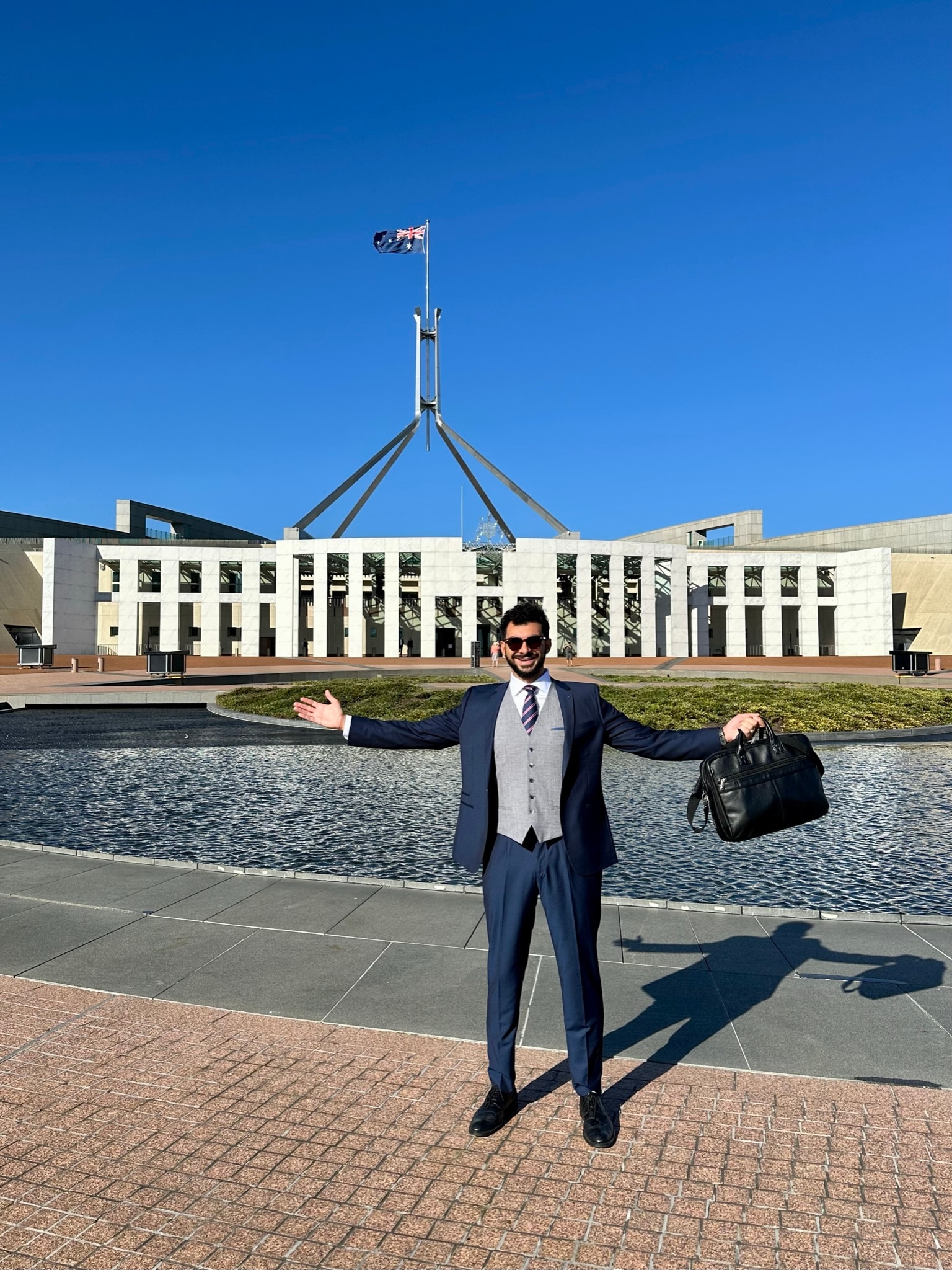 Man holding a bag with arms outstretched standing in front of Parliament House building and lake.
