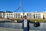 Man holding a bag with arms outstretched standing in front of Parliament House building and lake.