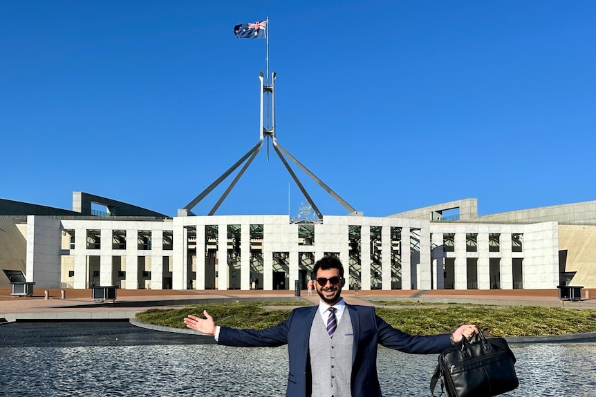 Man holding a bag with arms outstretched standing in front of Parliament House building and lake.