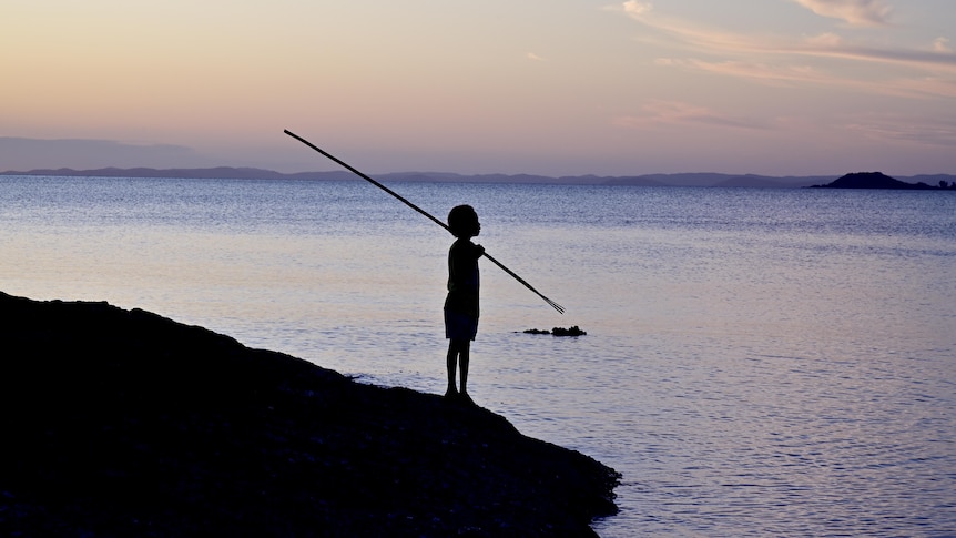 Silhouette of a small boy carrying a traditional fishing spear standing on the edge of the ocean looking at the horizon