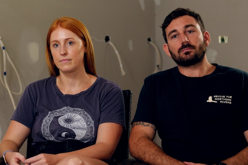 A woman with red hair and a man with a bear sitting in a room with wires exposed on the walls behind.