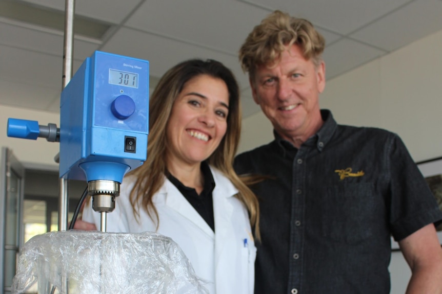 A smiling woman with long hair and a lab coat and a man in the black shirt in front of machinery and honey skin products.