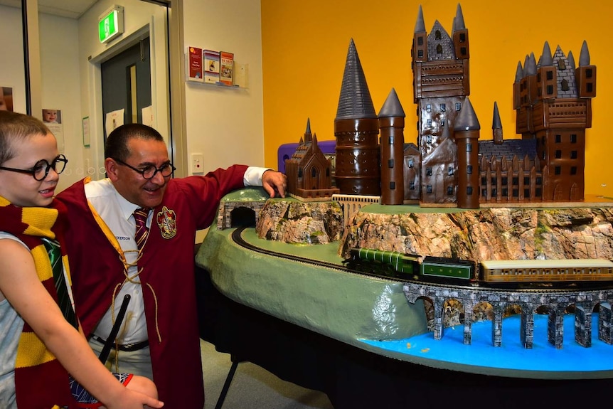 Middle-aged man and small boy dressed in Harry Potter costumes look at elaborate cake of Hogwards
