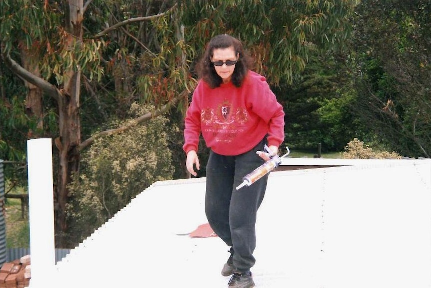 A woman in a red jumper and trackpants stands on a corrugated iron roof with a glue gun in her right hand.