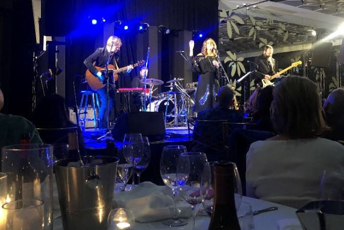 A woman stands on stage singing with two guitarist and a drummer as people watch on from their dinner table