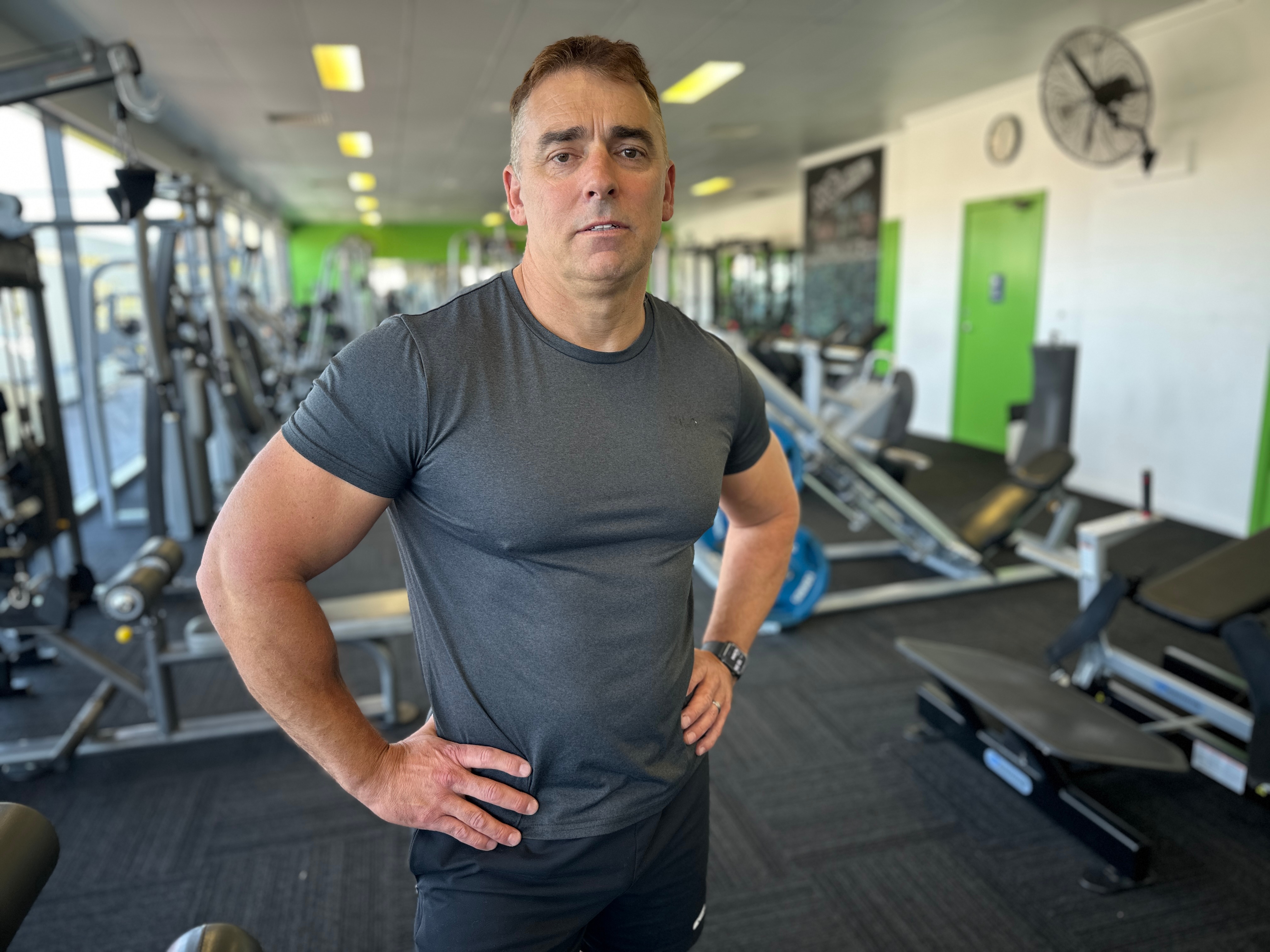 A muscly man in a blue t-shirt stands in front of a gym equipment