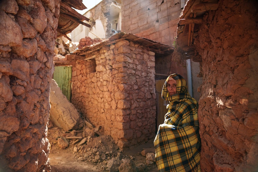 A person wearing a checkered scarf stands among rubble and leans against a stone structure.