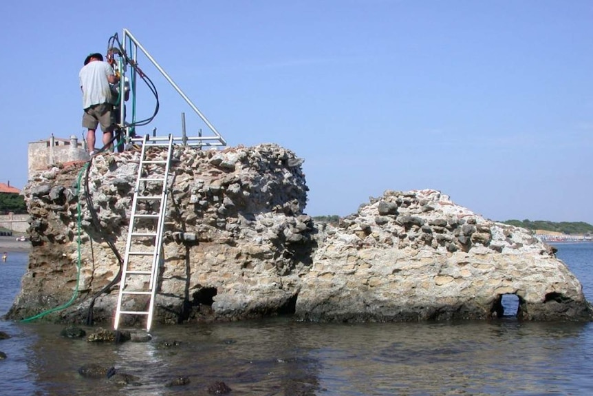 Man stands on top of a concrete wall in the sea collecting samples.