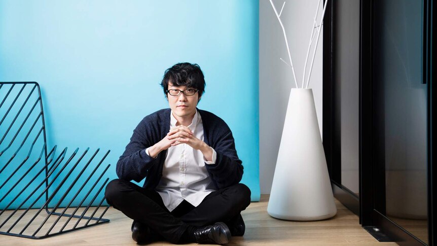 Man wearing white shirt, black pants, dark blue cardigan and black-rimmed glasses, sits on wooden floor in front of blue wall.