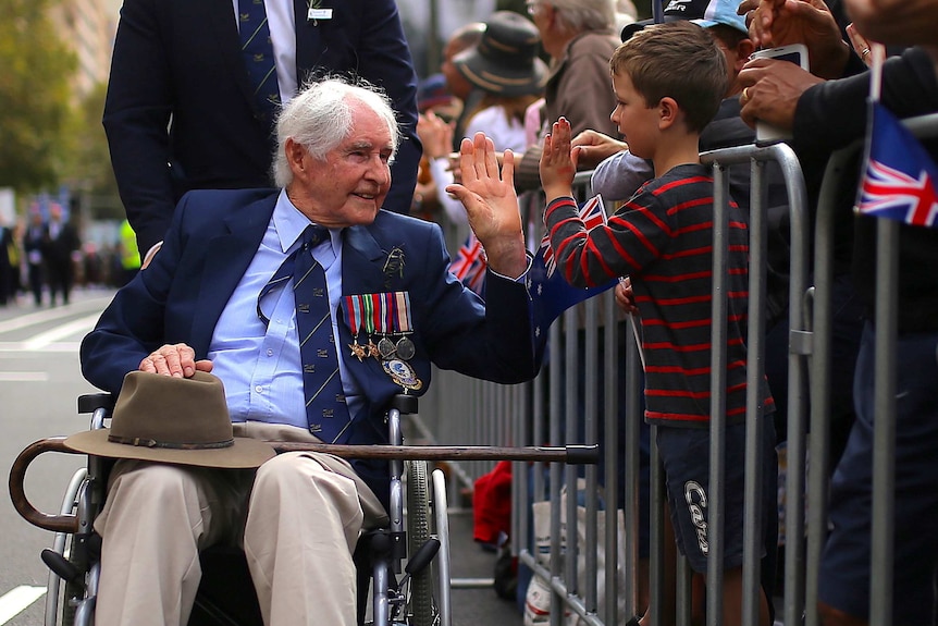 A war veteran wearing a suit and war medals is wheeled in a wheelchair and high fives a child through a road barrier